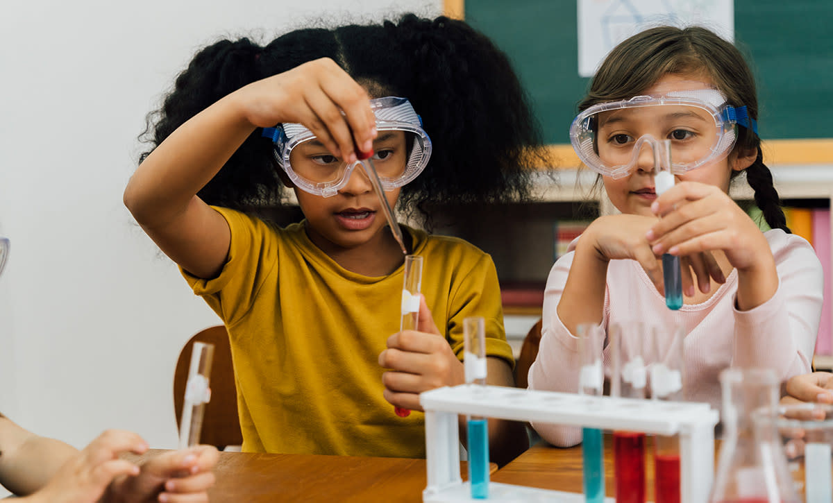 Two girls wearing safety goggles sitting at a table mixing liquids in vials during a chemistry lesson in school