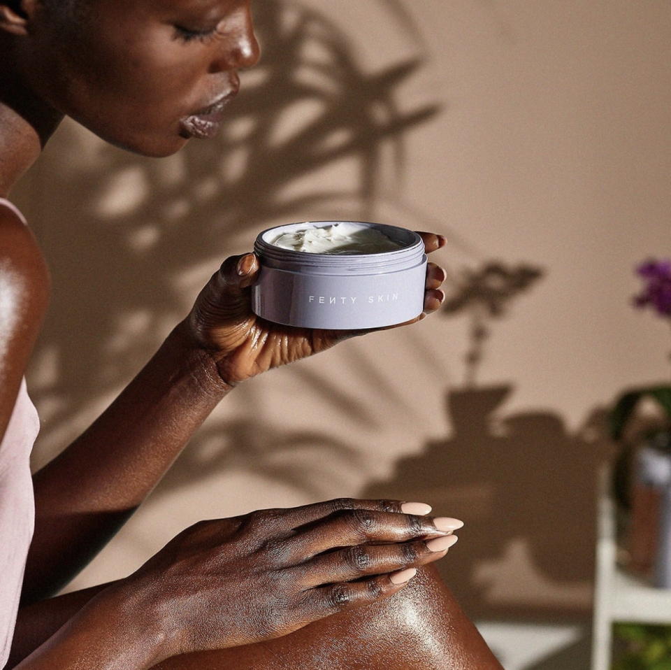 black model sitting applying fenty skin body butter from purple container on legs