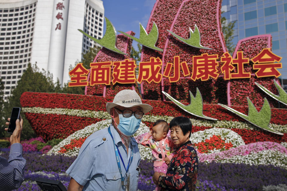 A man wearing a face mask to help curb the spread of the coronavirus walks by a woman with a child posing for a souvenir photo near a floral decoration baring the words "Fully built a well-off society" for celebrating the up-coming National Day in Beijing, Wednesday, Sept. 30, 2020. Even though the spread of COVID-19 has been all but eradicated in China, the pandemic is still surging across the globe with ever rising death toll. (AP Photo/Andy Wong)