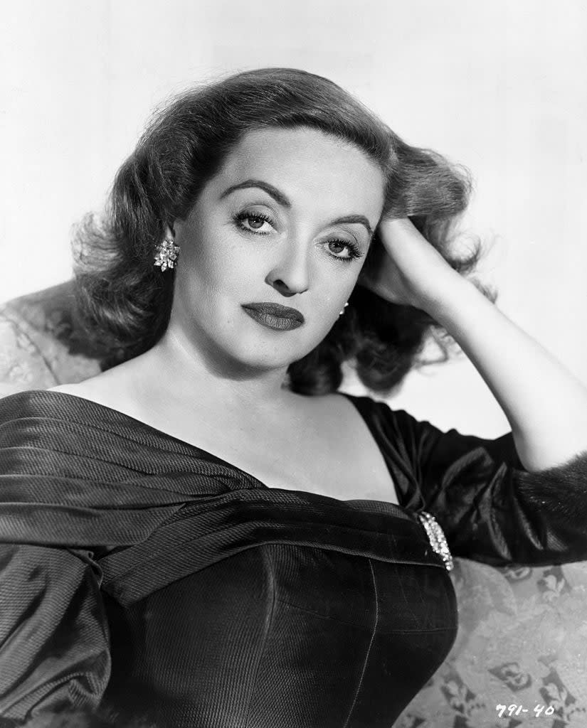 Bette Davis poses with her hand resting on her head, wearing an elegant off-shoulder dress adorned with a brooch, and earrings