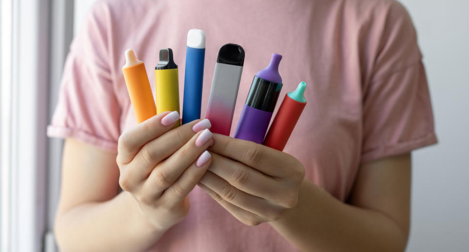 A young girl in a pink top holds up a selection of colourful vapes.