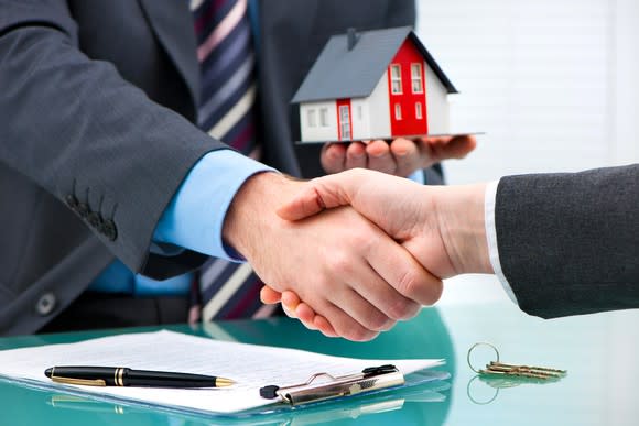 Two businessmen shaking hands after signing mortgage paperwork, with one holding a miniature house in his left hand.