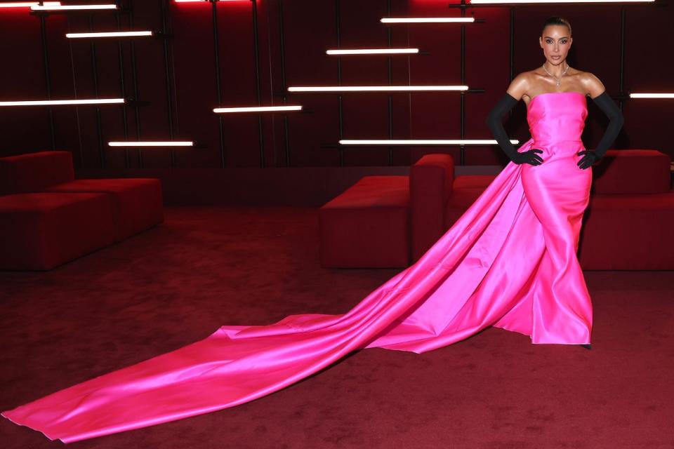 Kim Kardashian stands in a pink dress with black gloves in front of a red background.