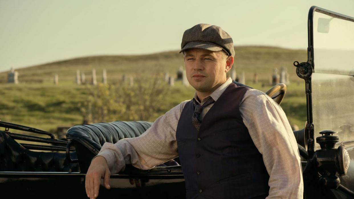 leonardo dicaprio in character for killers of the flower moon leans back against a 1920s car, he wears a suit vest, collared shirt, tie and cap