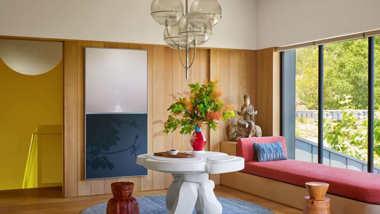 sitting area has a built in oak window seat with muted vermillion cushion, a carved limestone center table, a stool left and right of table, 4 globe pendant, an organic shaped blueish rug, a sculpture and artwork