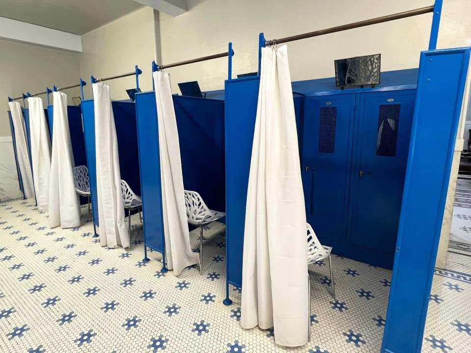 A changing area with blue stalls, blue lockers, and white chairs covered by sliding white curtains. The floor has blue-and-white tiling.