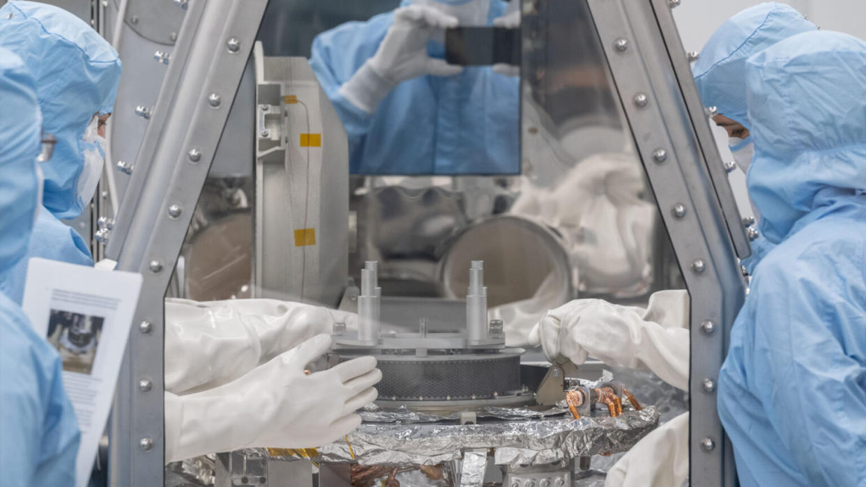  Technicians in blue 'bunny suits' manipulate a metallic object inside a clean room. 