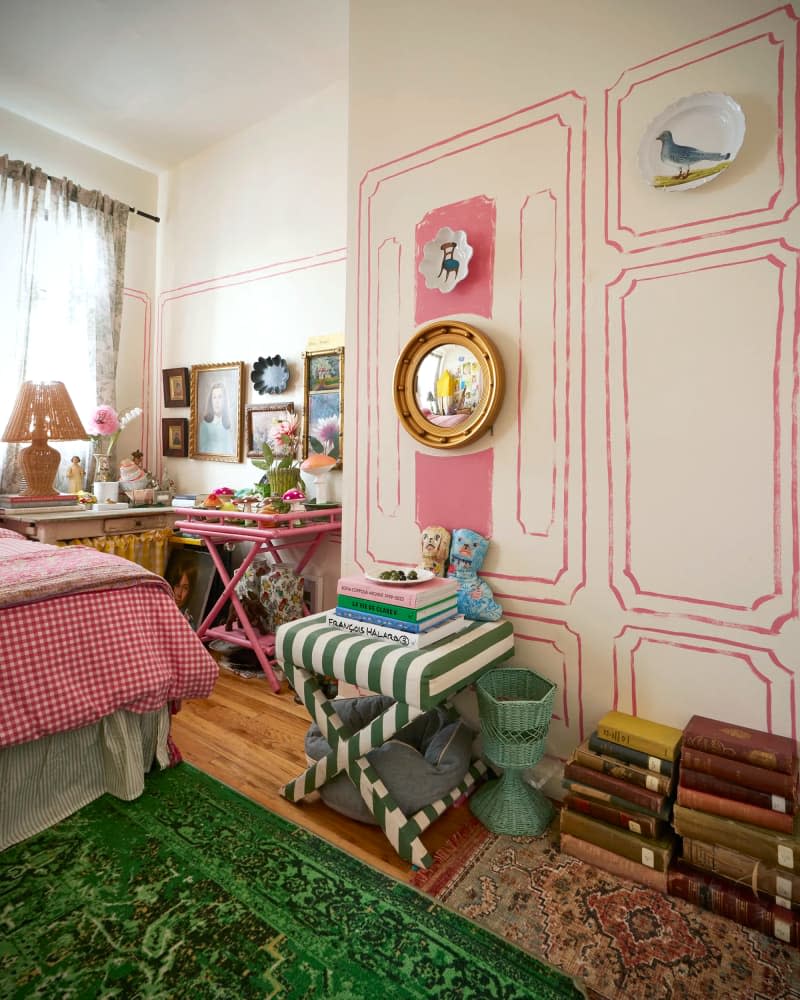 Mirror and plates on white walls with pink trompe l'oeil detail above green striped stool and stacked books in bedroom with green area rug.