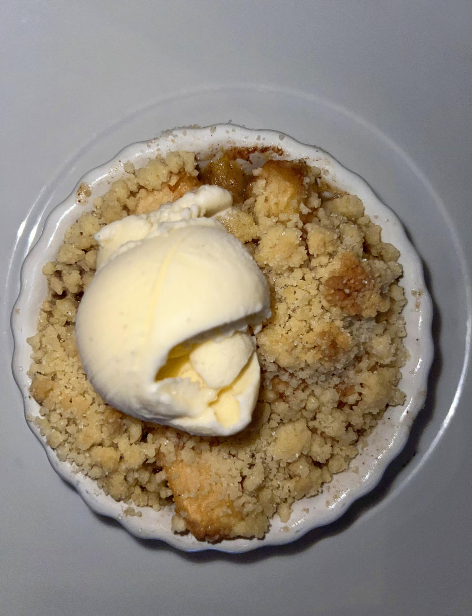 A serving of apple crumble in a white dish, topped with a scoop of vanilla ice cream