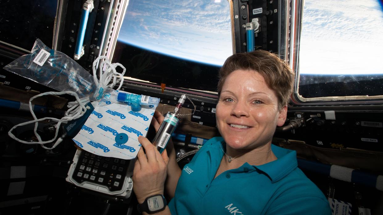  anne mcclain floats in a space station in front of two big windows facing earth. in her hands is a tangle of bags, sampling cubes and wires for a science experiment 