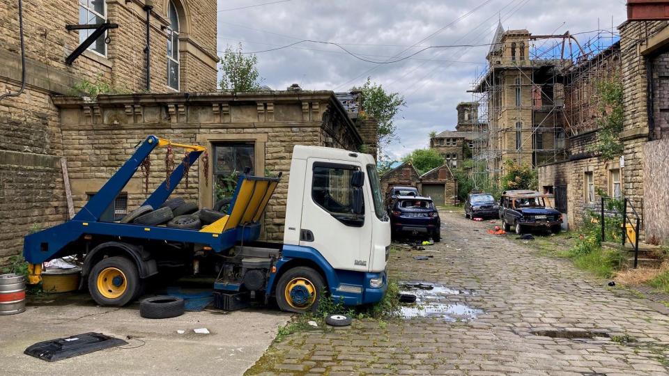 An abandoned skip truck and burnt out vehicles line a cobbled street inside the mill complex