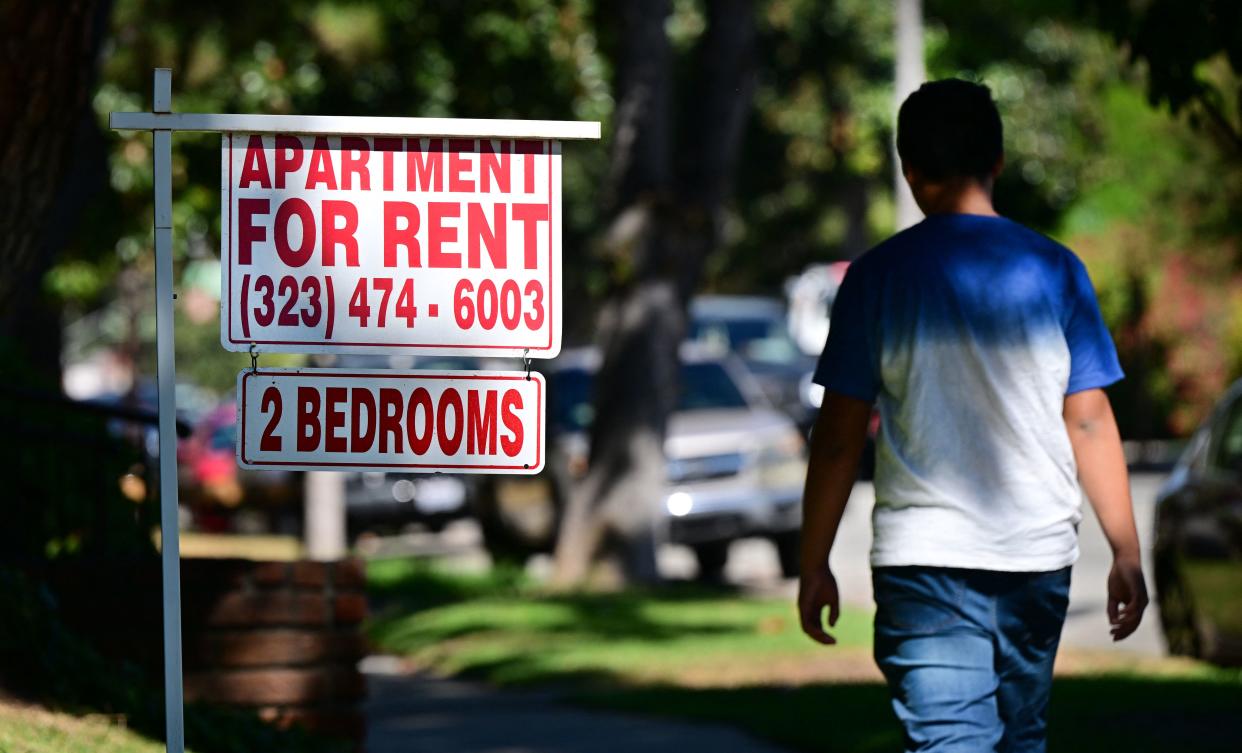 A man walks down a leafy avenue past a sign that says: Apartment for Rent (323) 474-6003, 2 Bedrooms.