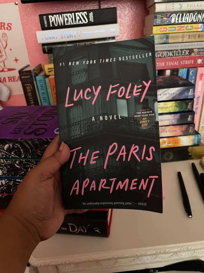 A person holds the book "The Paris Apartment" by Lucy Foley, a New York Times bestseller, in front of a bookshelf filled with various other books