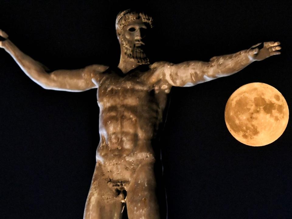 An ancient Greek statue of Poseidon, naked with arms outspread, against a dark sky with a full moon, in Athens, Greece on August 11, 2022.