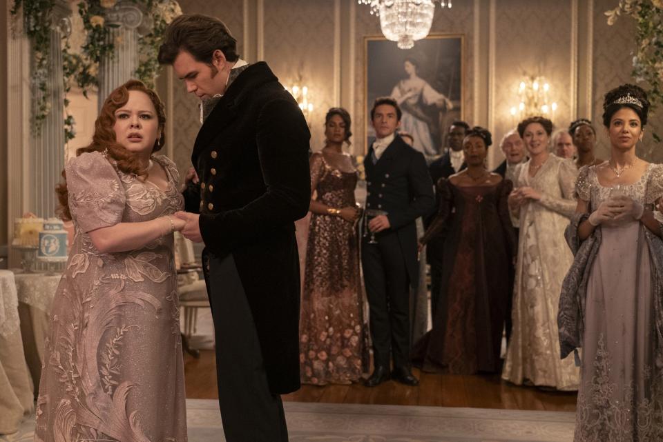 Nicola Coughlan and Luke Thompson in elegant period attire, in a grand room with a group of formally dressed individuals from the "Bridgerton" series