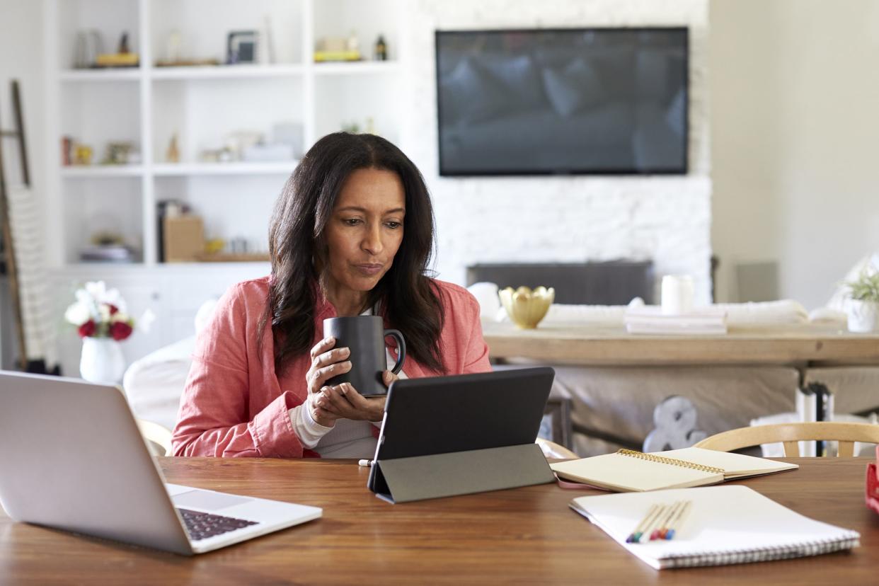 middle aged woman sitting at a table reading using a tablet computer, holding a cup