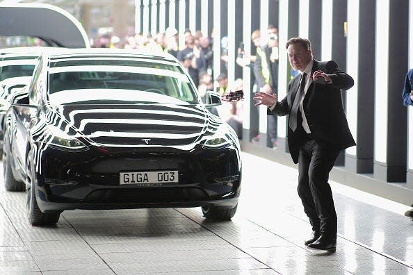 Elon Musk wearing a suit and standing next to a black Tesla at a car manufacturing plant.