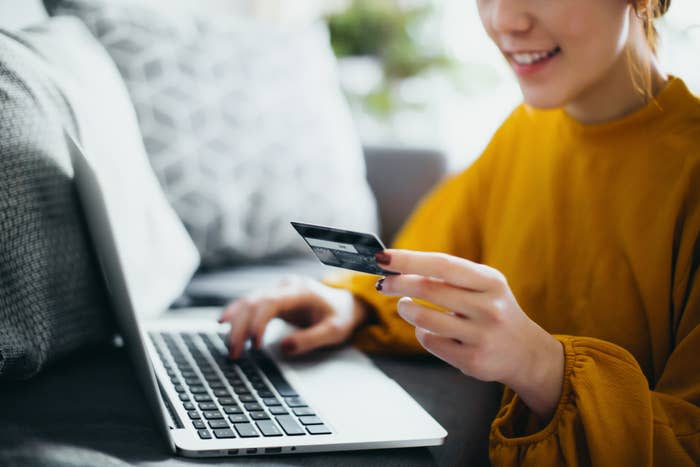 A smiling person in a long-sleeved shirt uses a credit card while typing on a laptop, indicating online shopping or managing finances