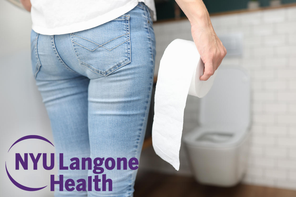 Woman standing with toilet paper roll approaching a toilet.