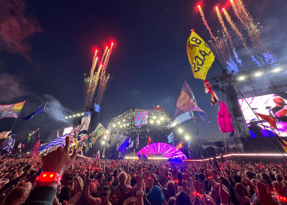 Large crowd at an outdoor music festival at night, with colorful flags and bright fireworks lighting up the sky above the stage