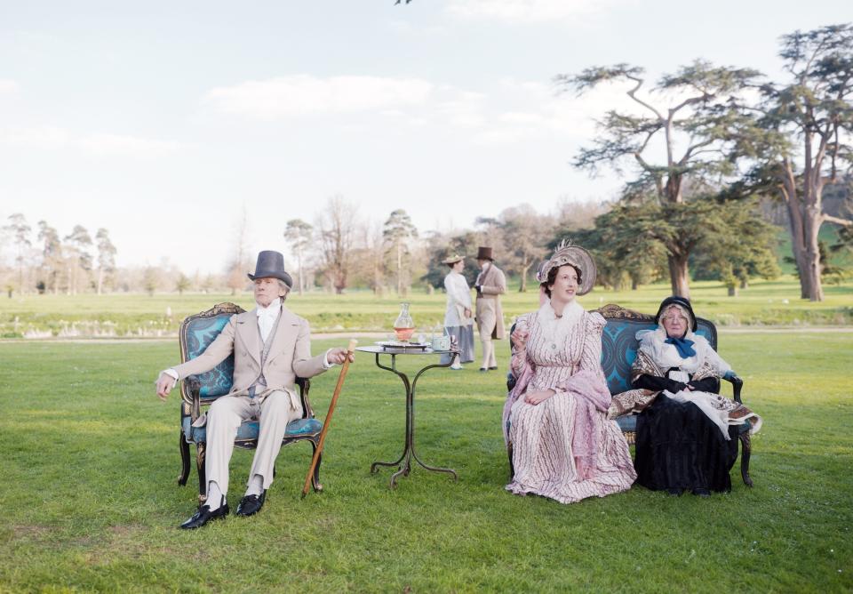 3 characters at the forefront sitting on fancy chairs outside with side table holding their drinks dressed in 19th-century clothes