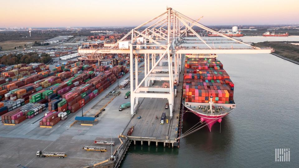 Image shows container cranes, container stacks, container vessel and waterway.