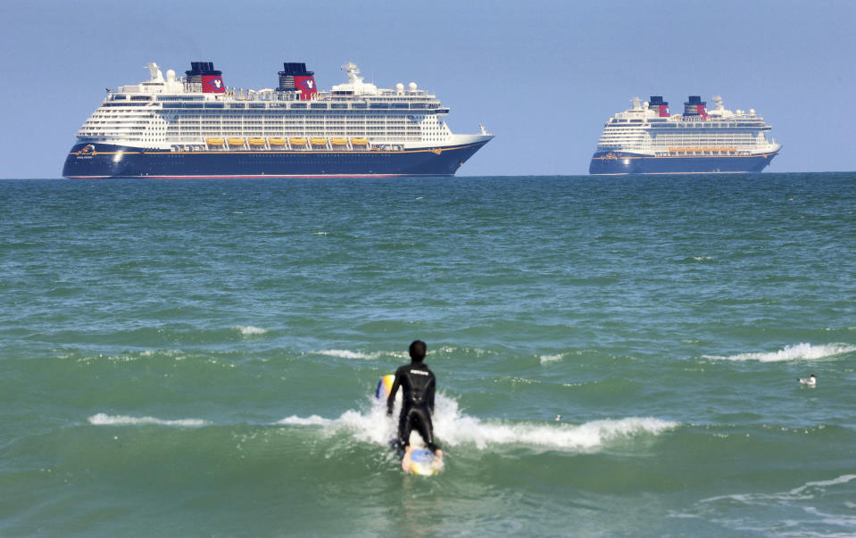 FILE - In this March 24, 2021 file photo, a surfer eyes the Disney Cruise Line ships "Fantasy, " left, and "Dream" on the horizon as they sit stationary off of Cocoa Beach, Fla., in this view from Lori Wilson Park. Florida is threatening to sue the federal government if it doesn't lift its pandemic ban on cruise lines using U.S. ports. Gov. Ron DeSantis and state Attorney General Ashley Moody said Friday, March 26, that the state is being harmed economically by the industry's U.S. shutdown. (Joe Burbank/Orlando Sentinel via AP, File)