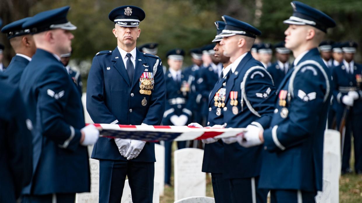  uniformed military personnel surround a stretched-out american flag within view of gravestones in arlington cemetery. far in the background are military members standing  