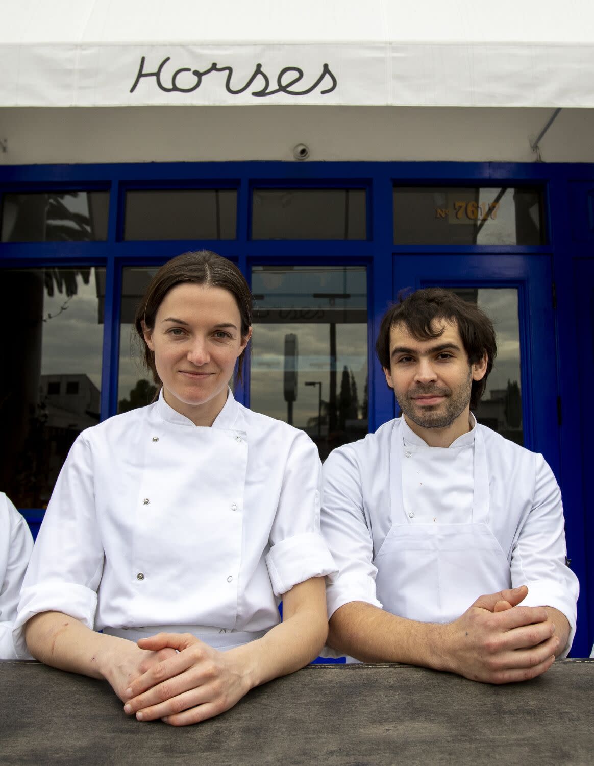 Two people in white chef coats are framed waist up standing and looking at the camera.