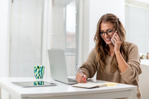 Young woman sitting at a laptop, taking notes, while on a cellphone.