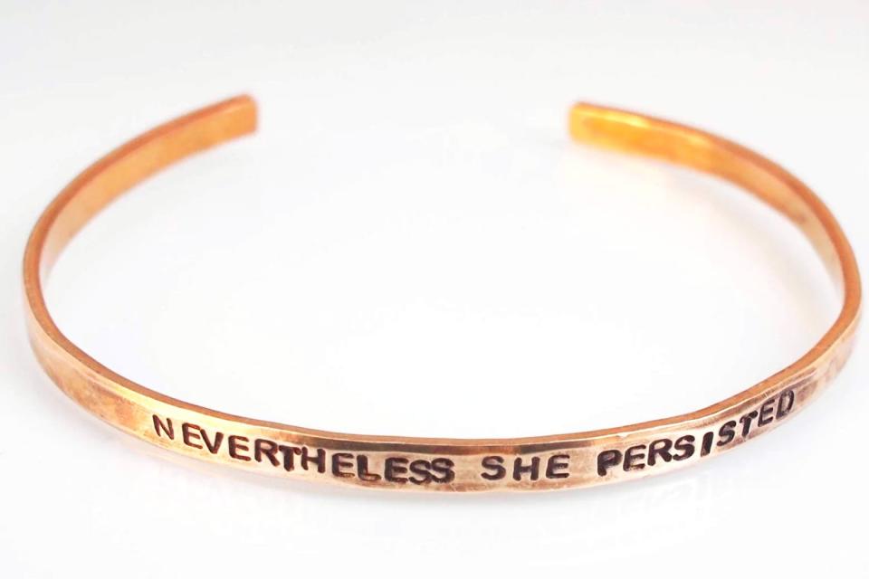 A gold bracelet embellished with the quote "Nevertheless She Persisted."