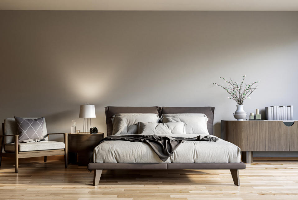 Modern minimalist bedroom featuring a bed with beige linens, a nightstand with a lamp, a comfortable chair, a vase with branches on a sideboard, and stacked books