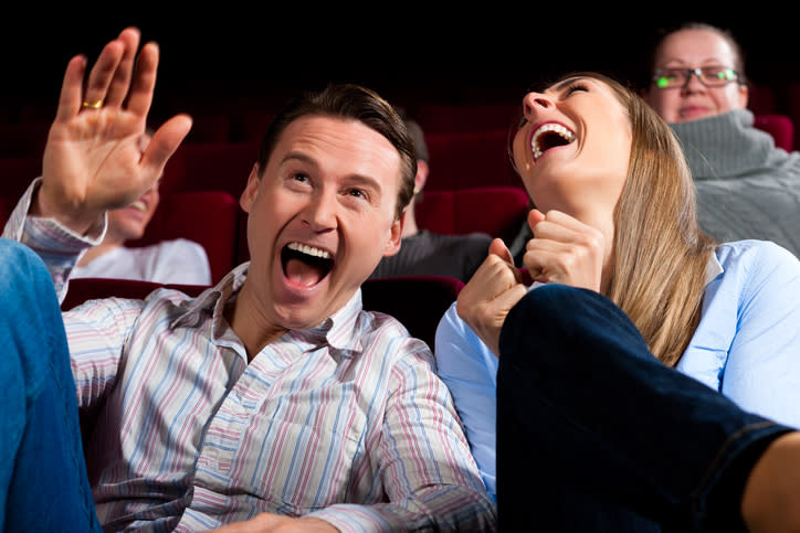 A man, wearing a casual shirt, and a woman, wearing a blouse, laugh heartily while seated in a theater. They are surrounded by other laughing people