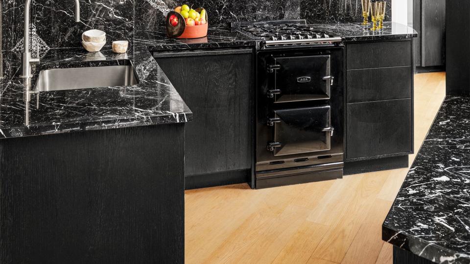 a kitchen has black lower cabinets topped with white veined black marble and upper cabinets with glass fronts showing glassware and dishes, stainless steel sink, and planked oak flooring