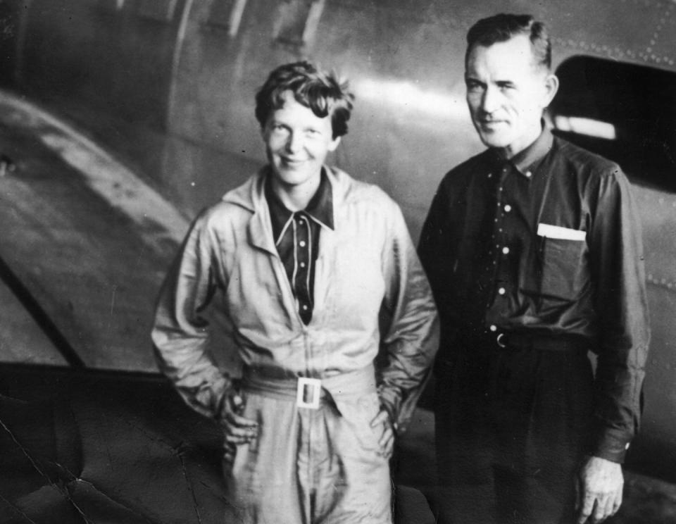 amelia earhart and fred noonan stand near a plane and smiles, she wears a jumpsuit with her hands on her hips, he wears a collared shirt and pants