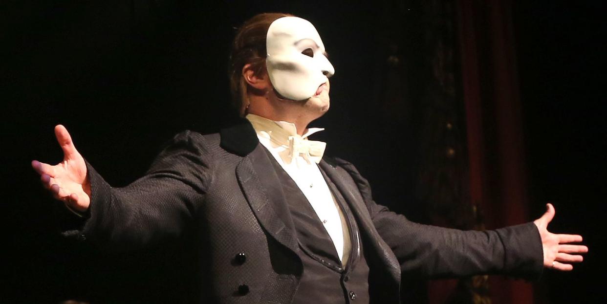 ben crawford dressed as the phantom of the opera, spreading his arms toward the audience while standing on a stage, with other actors standing behind him