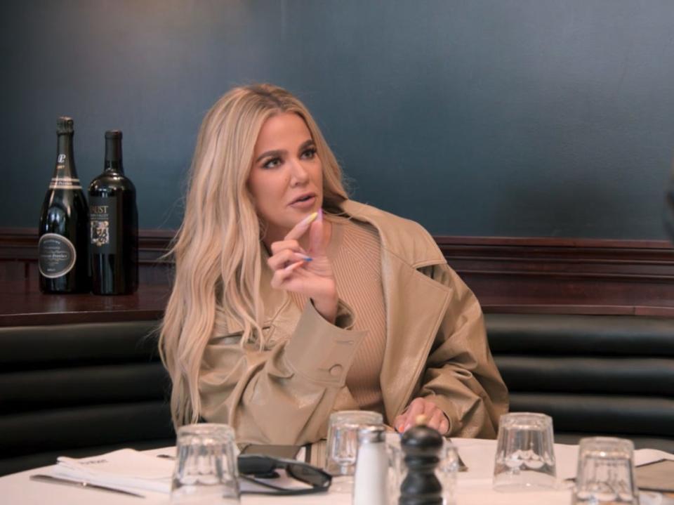 khloé kardashian sitting at a table, gesturing with her fingers very close together as if to demonstrate the thinness of something