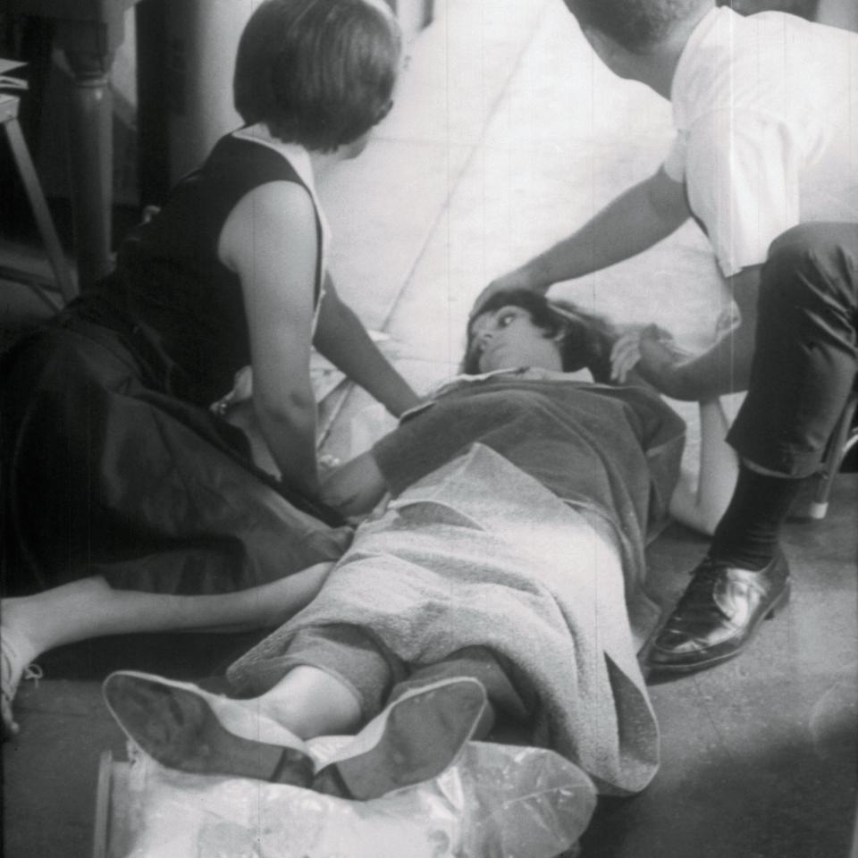 Three people are in a corridor. One is lying on the ground, covered with a blanket, being attended to by two others who appear concerned