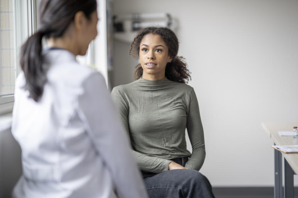 A young female patient sits casually with her doctor as they discuss her mental health.  She is seated in a chair in front of her doctor as they talk about her needs.  The doctor is wearing a white lab coat and has her back to the camera as the two discuss possible plans of care to navigate the woman's struggles.