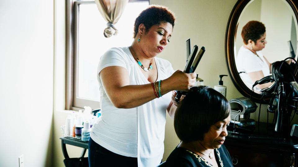 A hairdresser dressed in a white t shirt and dark pants can be seen straightening a woman's hair at a salon. The hairdresser has a concentrated look on her face as she is straightening the hair, holding the straighteners in one hand and scissors in the other. The client is looking down. A mirror and a bunch of hair tools on a surface can be seen in the background