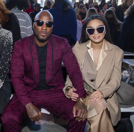 A bald Black man in a maroon suit and sunglasses sitting with an Asian woman with long hair in a beige suit and sunglasses