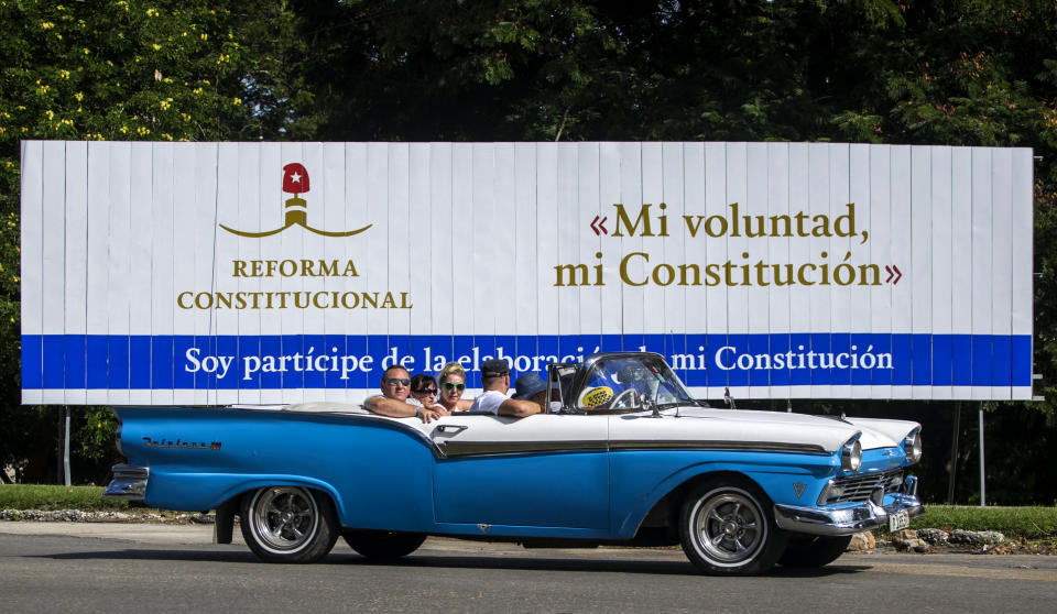 FILE - In this Oct. 2, 2018 file photo, tourists take a joy ride in a vintage convertible car, past a billboard promoting constitutional reform with the Spanish messages: "My will, my constitution," right, and "I'm a participant in the making of my constitution" in Havana, Cuba. In the midst of a regional crisis over Venezuela and tough economic straits, the Cuban government is about to launch a sweeping makeover of its centrally planned, single-party system with dozens of new laws that could reshape everything from criminal justice to the market economy. (AP Photo/Desmond Boylan, File)