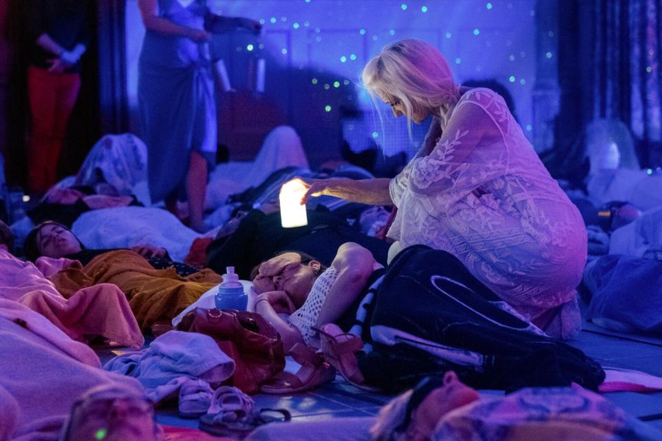 A woman in a flowing white dress perches over guests on the floor in a darkened room during a sound bath.