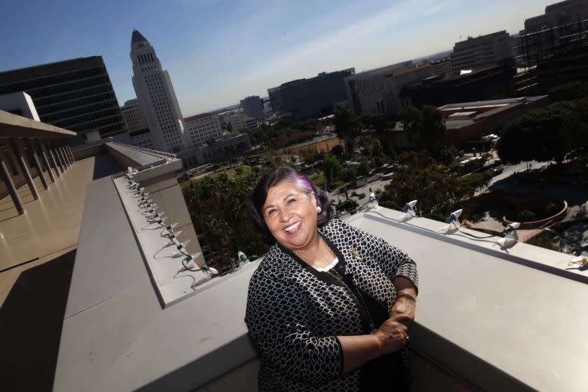 Gloria Molina poses for a portrait in sunlight, smiling. The LA City Hall is in the backgroung.