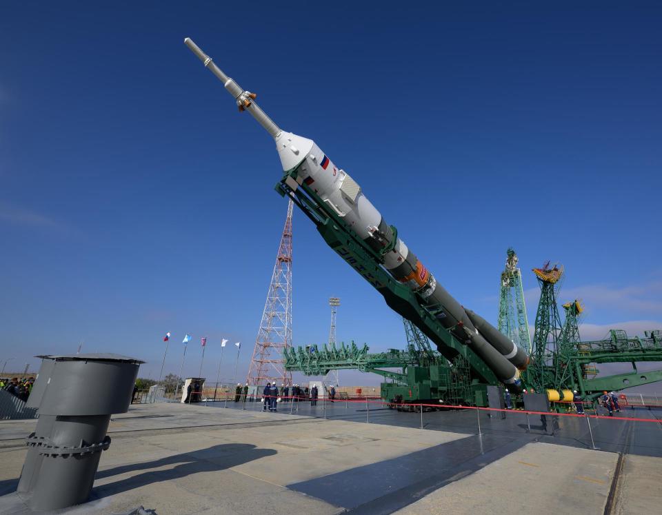 A green/grey soyuz rocket with a white pointy top is laying diagonally as a green support arm that stretches the rocket's height lifts the vehicle vertical.