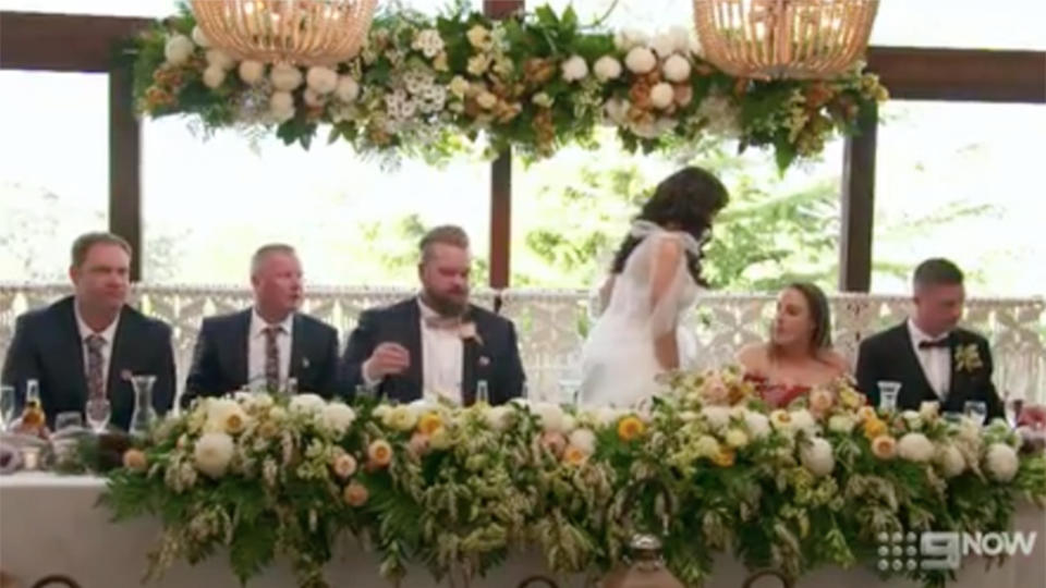 Bride Poppy leave table in wedding dress guests and groom watch