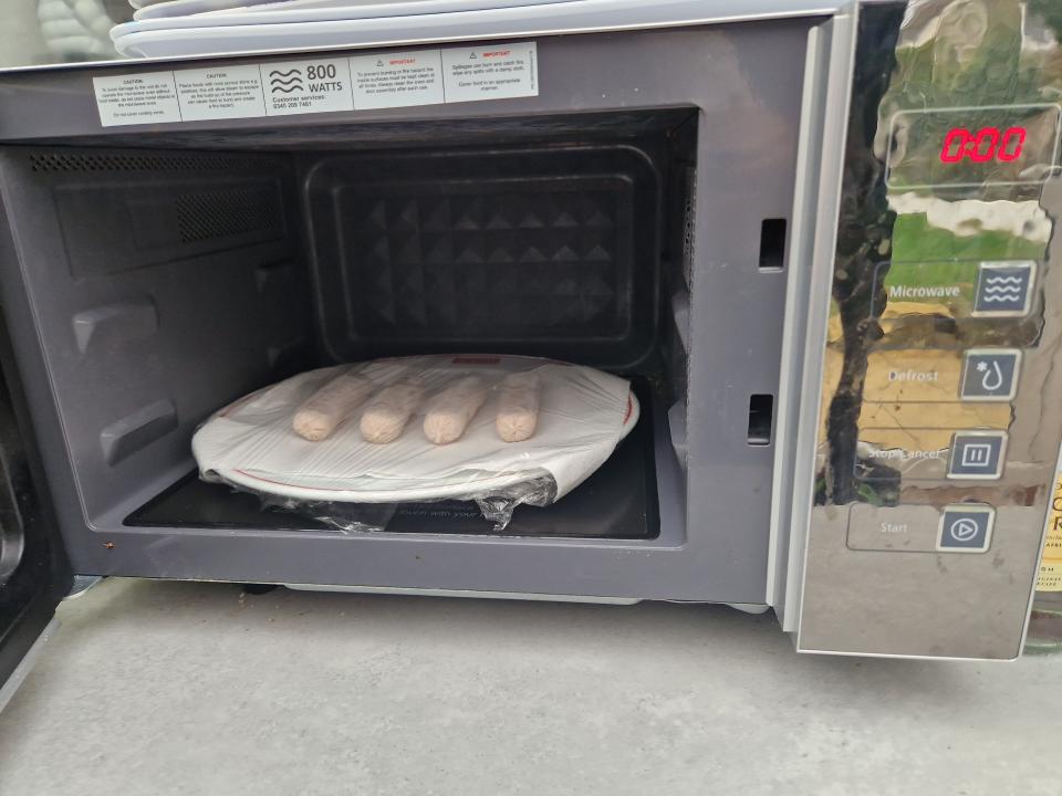 four sausage links on a plate covered in plastic wrap in a microwave