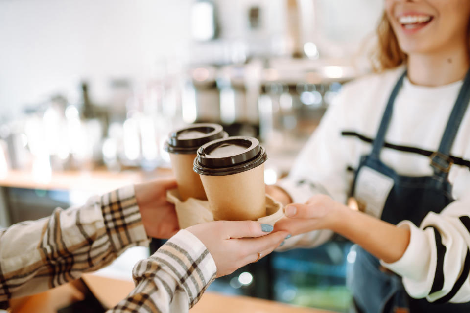 Barista in an apron handing two coffee cups to a customer's outstretched hands