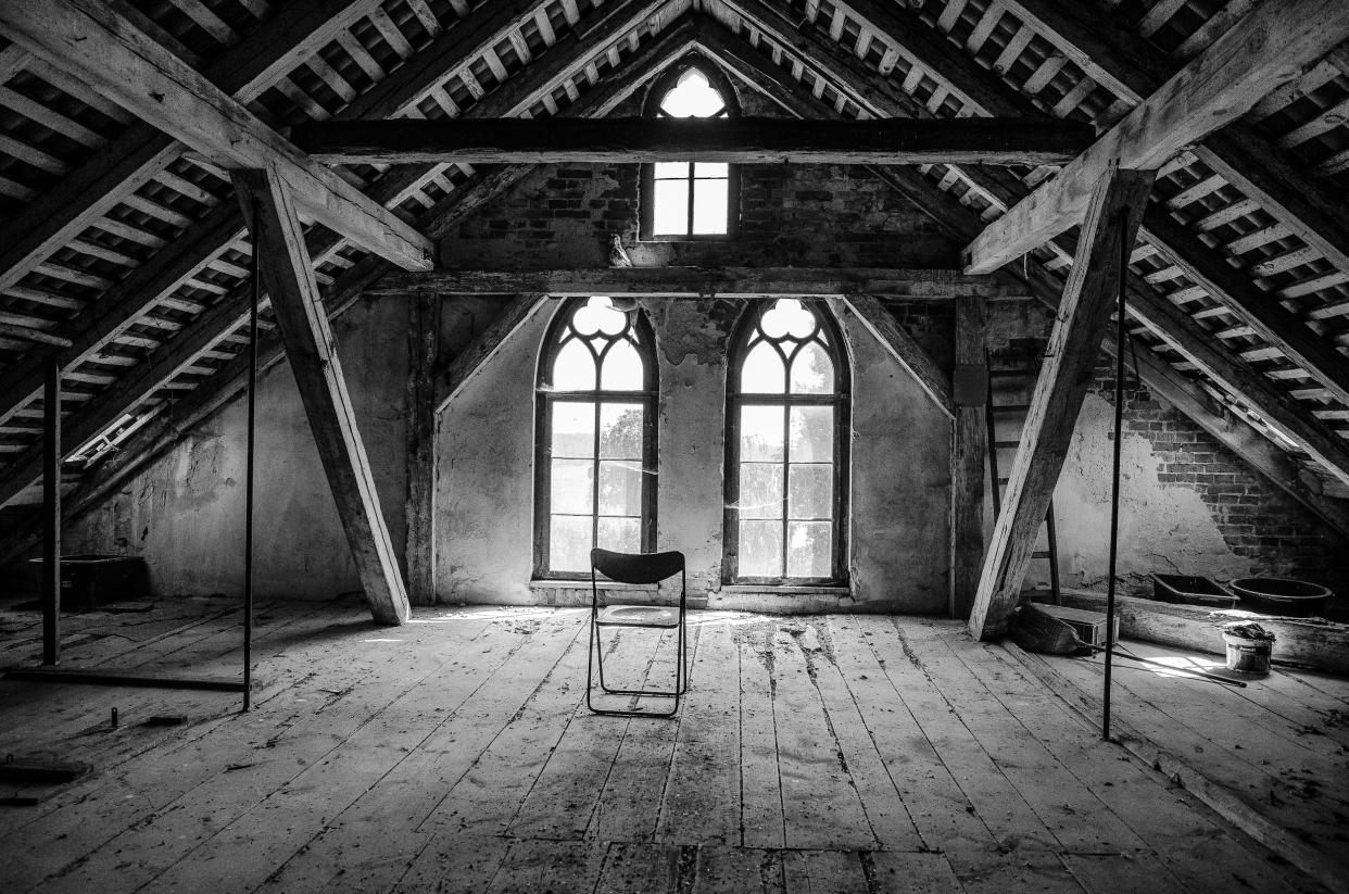  Black and white photo of an empty wooden attic with a metal folding chair in the center. 