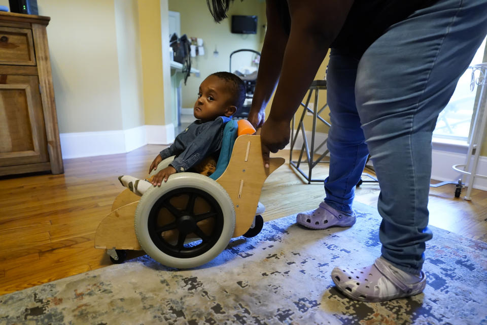 Crystal Jack assists her son Elijah Jack, 1, in his mobility chair, which was built as a donation by Tulane University students, at her home in New Roads, La., Thursday, Nov. 30, 2023. Tulane science and engineering students are making the second batch of mobility chairs for toddlers, that will eventually go to pediatric patients at Children's Hospital. Wheelchairs are expensive, and insurance won't cover the cost for children unless the child proves they can operate it independently. (AP Photo/Gerald Herbert)